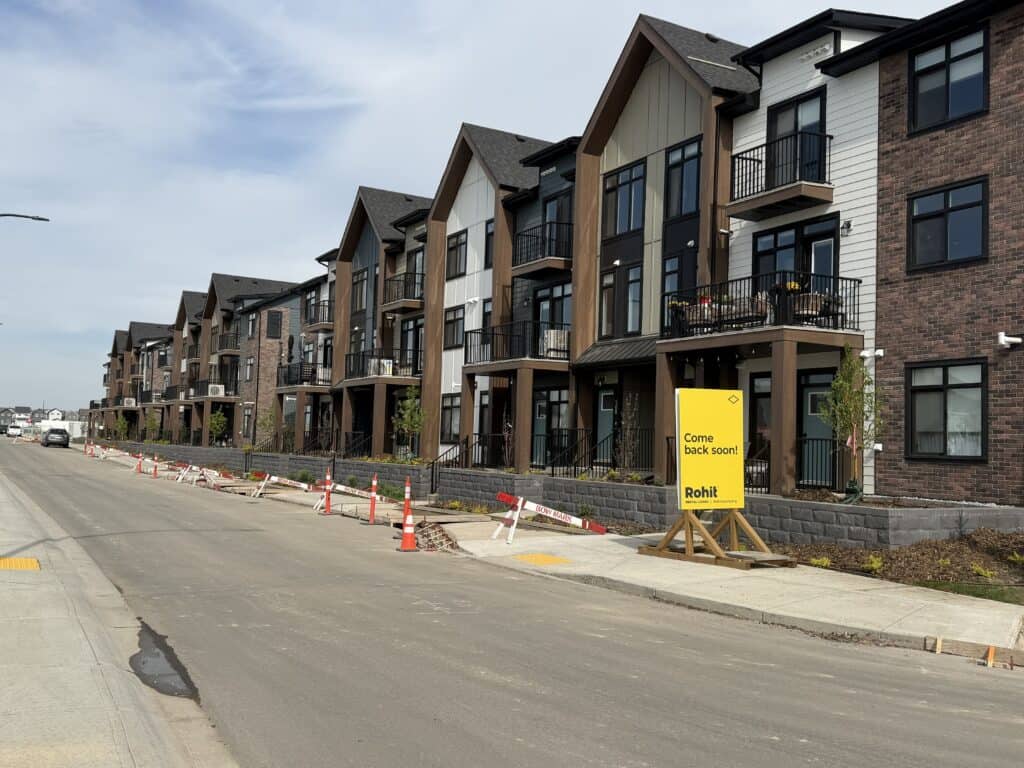 A modern multi-story residential building with balconies, under construction, along a street with construction cones and a "Come back soon!" sign.