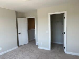 A well-lit bedroom with an open closet and an adjacent door leading to another room, featuring a freshly painted neutral wall colors and beige carpet flooring.